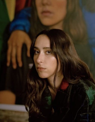Person with long dark hair seated indoors, wearing a patterned jacket, with a thoughtful expression.