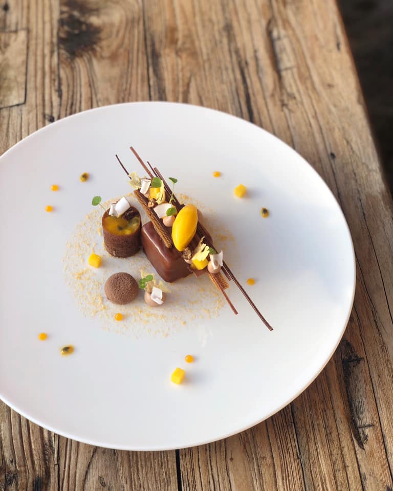 Gourmet dessert with chocolate mousse, mango sorbet, and garnishes on a white plate atop a rustic wooden table.