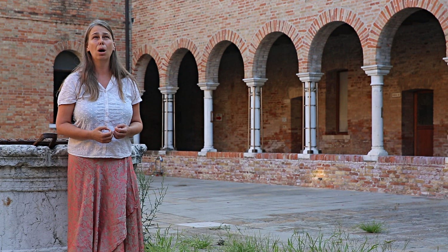 Woman standing in a historic courtyard with arched brick walls and stone columns, speaking or singing passionately.