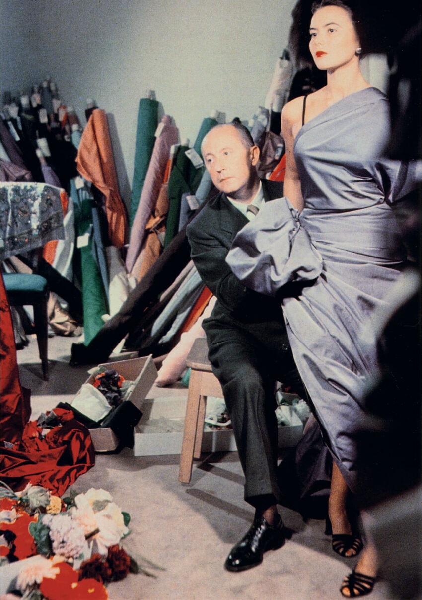 Designer adjusting a model's elegant blue gown in a workshop surrounded by vibrant fabric rolls and colorful flowers.