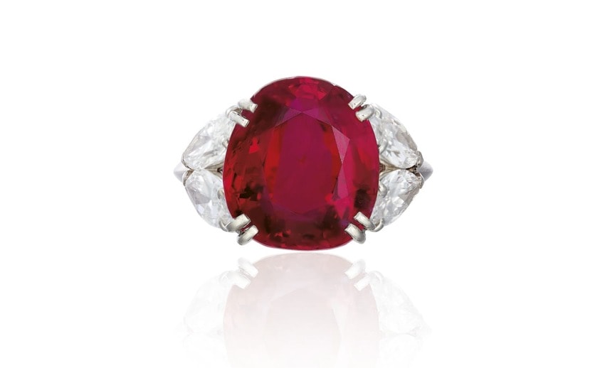 Oval red ruby ring with two pear-shaped diamonds on each side, set in a silver band, displayed on a white background.