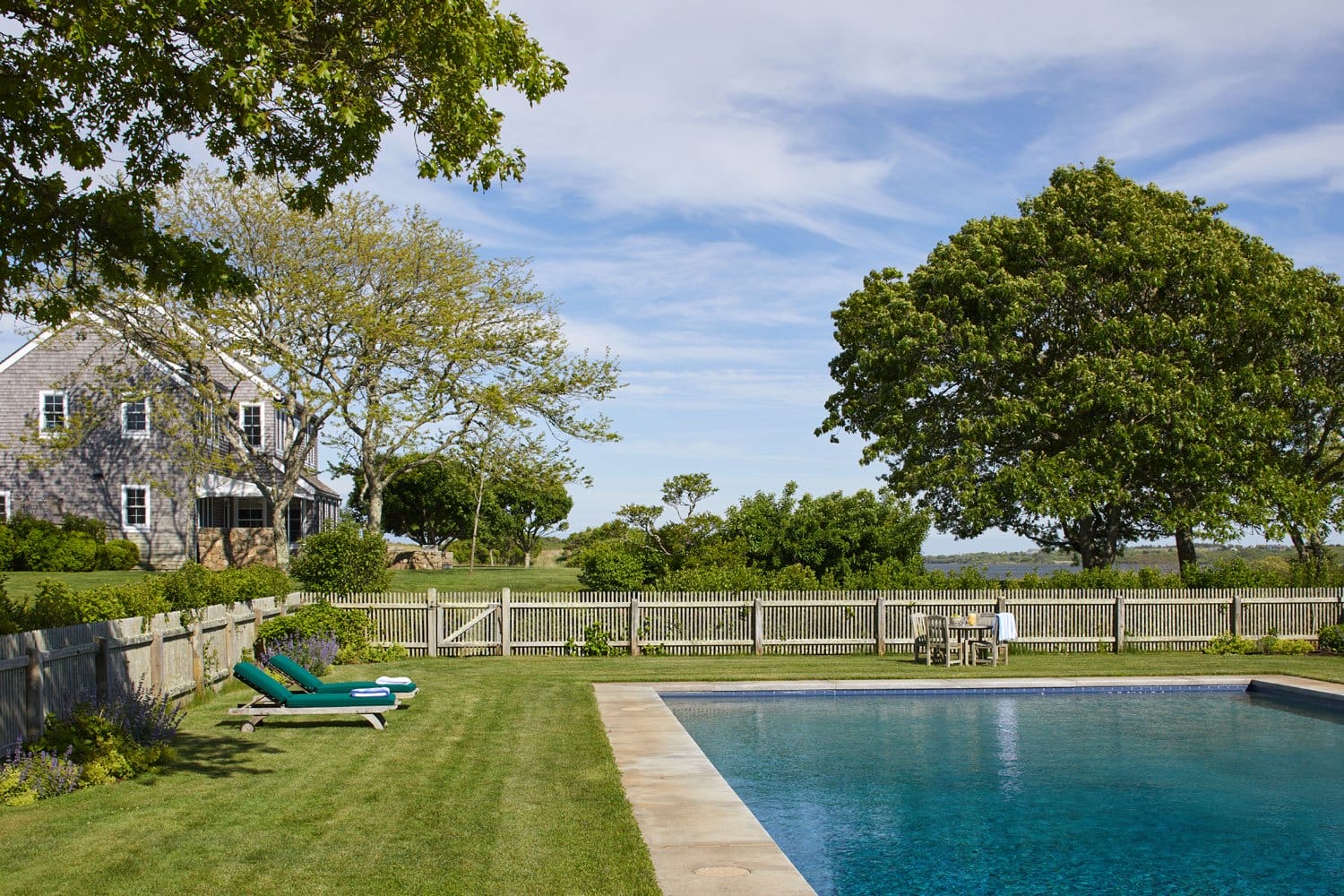 Backyard view of a traditional house with a pool, lawn chairs, trees, and a wooden fence under a blue sky.