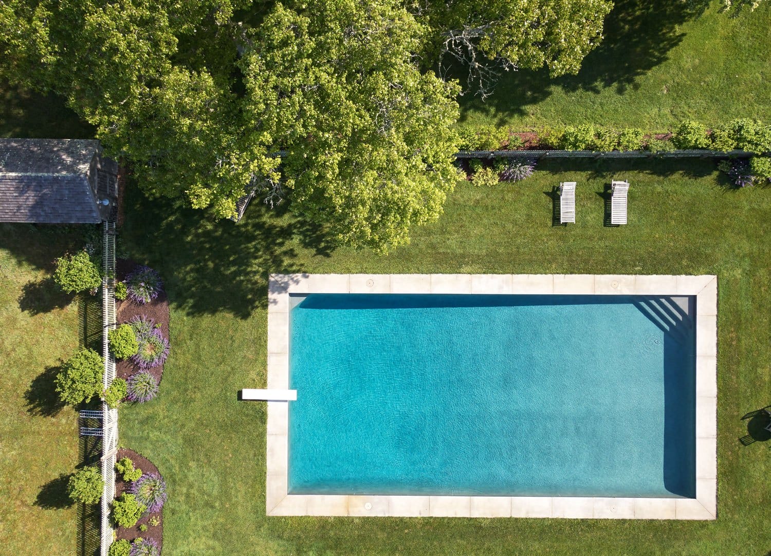 Aerial view of a rectangular swimming pool surrounded by green grass, trees, and two lounge chairs.