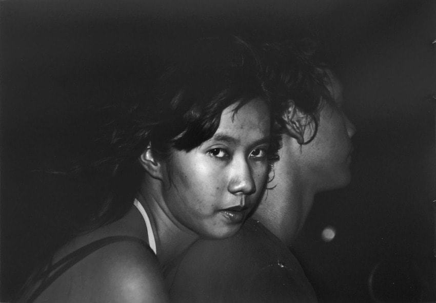 Woman looking at camera with another person facing away in a dimly lit black and white photograph.