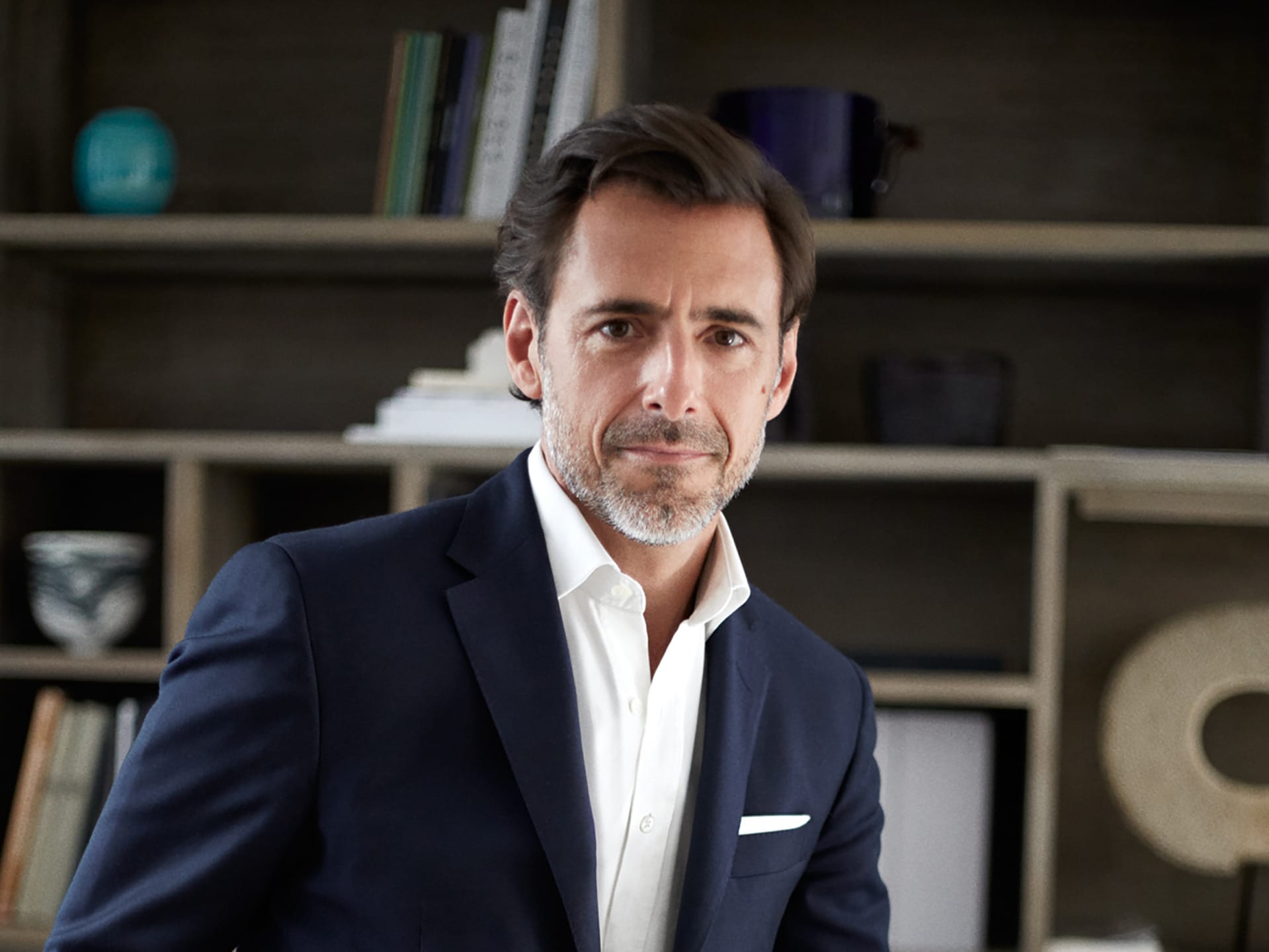 Man in a dark suit with a white shirt standing in front of a bookshelf filled with books and decorative items.