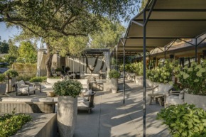 Outdoor patio with comfortable seating, shaded areas, and lush greenery under a clear sky.