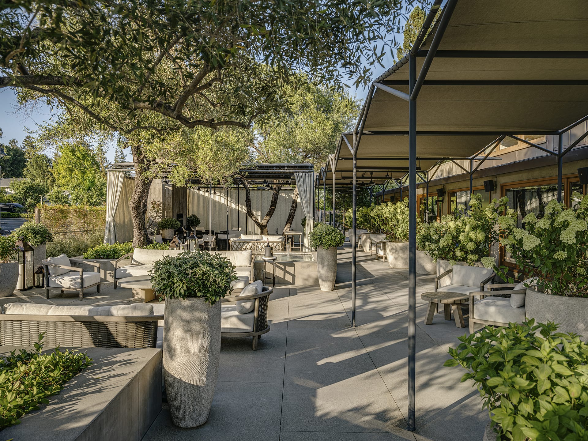 Outdoor patio with comfortable seating, shaded areas, and lush greenery under a clear sky.