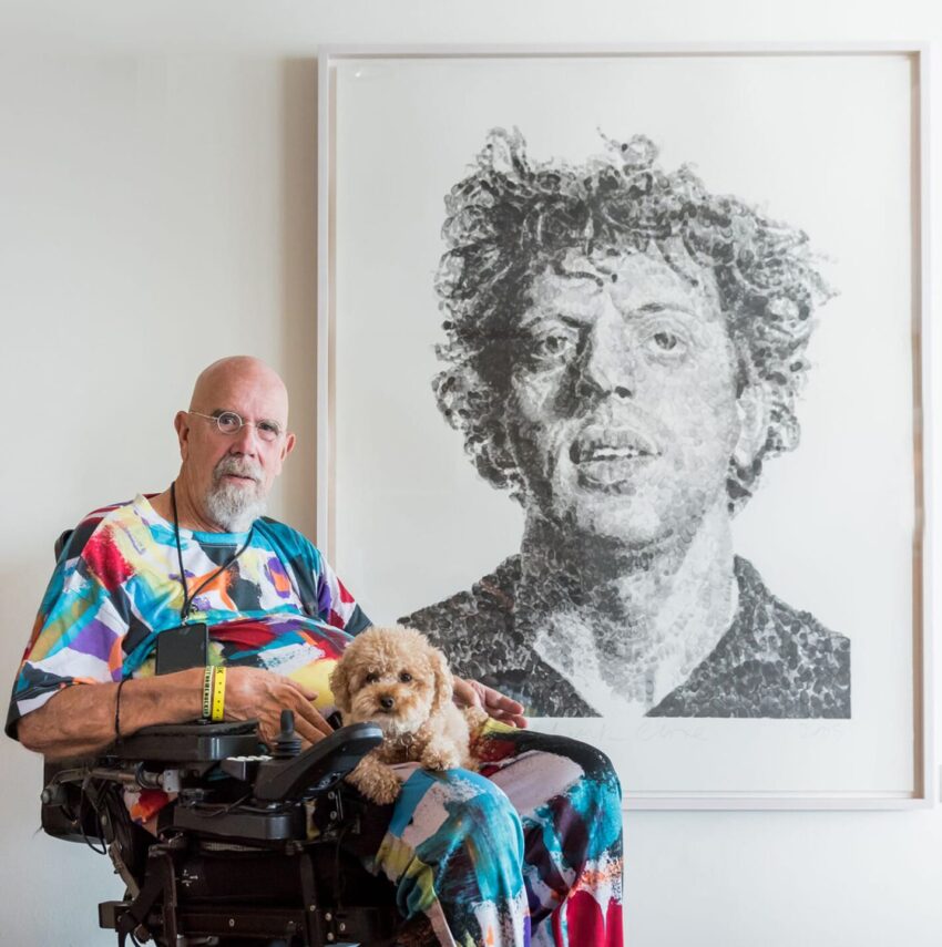 Man in a colorful shirt in a wheelchair holding a small dog, with a large portrait on the wall in the background.