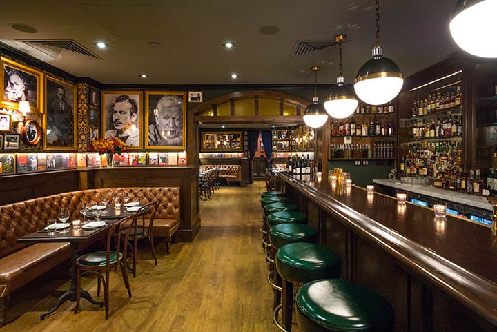 Cozy vintage-style restaurant interior with a wooden bar, green stools, framed photos on walls, and dim lighting.