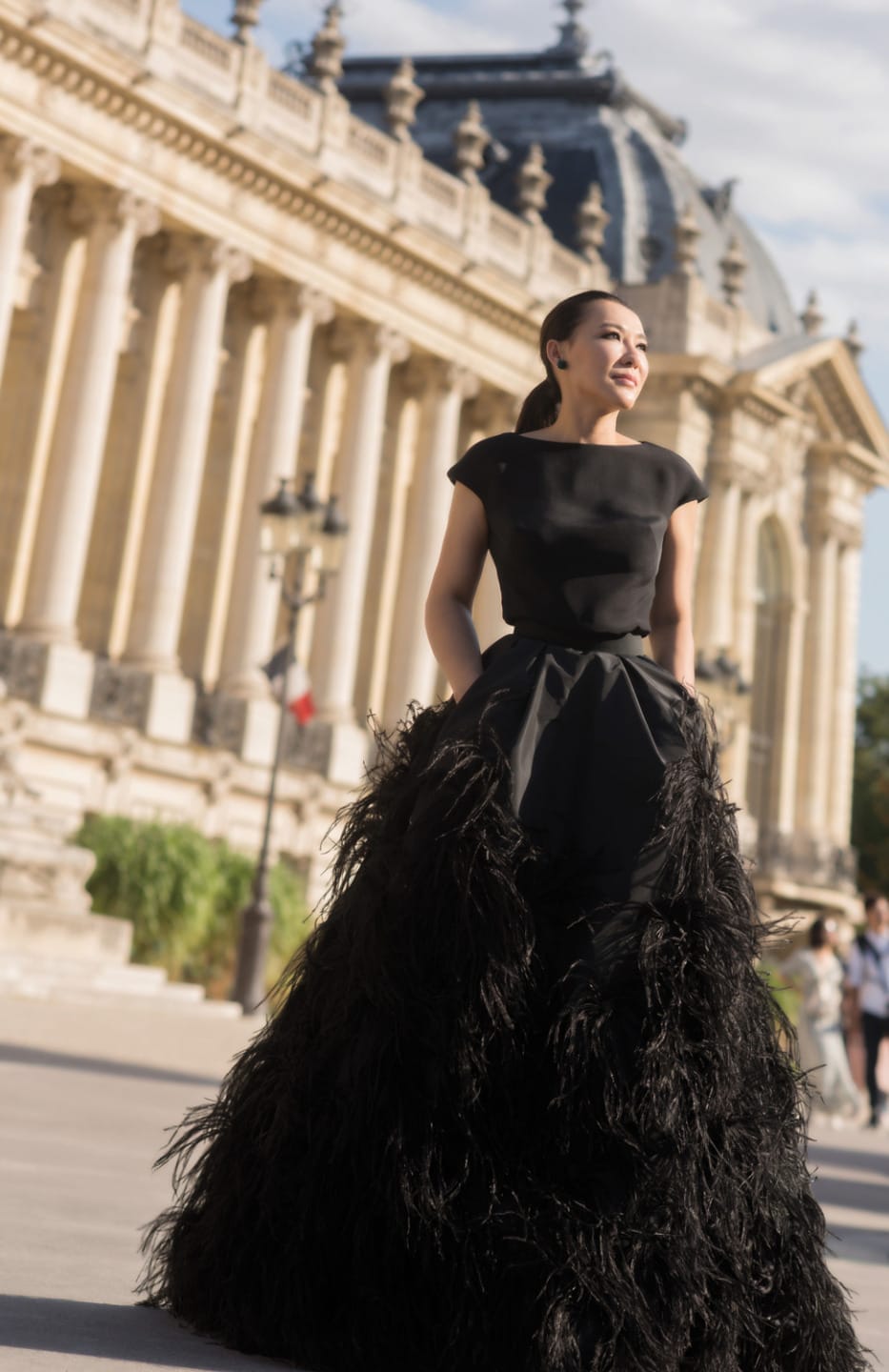 A woman in an elegant black gown stands near a historic building on a sunny day.