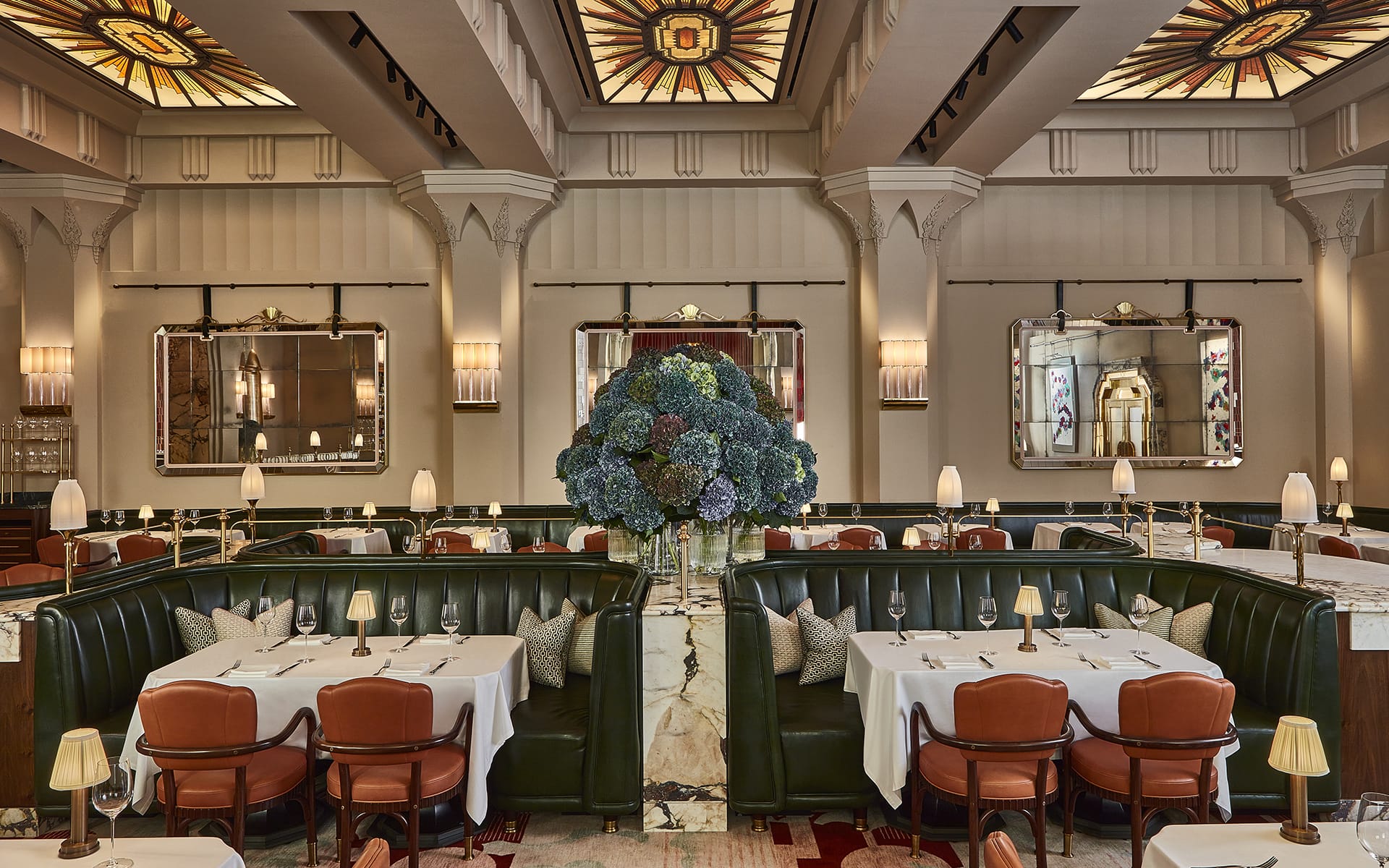 Elegant restaurant interior with plush seating, round tables set for dining, and large floral centerpiece under stained glass ceiling.