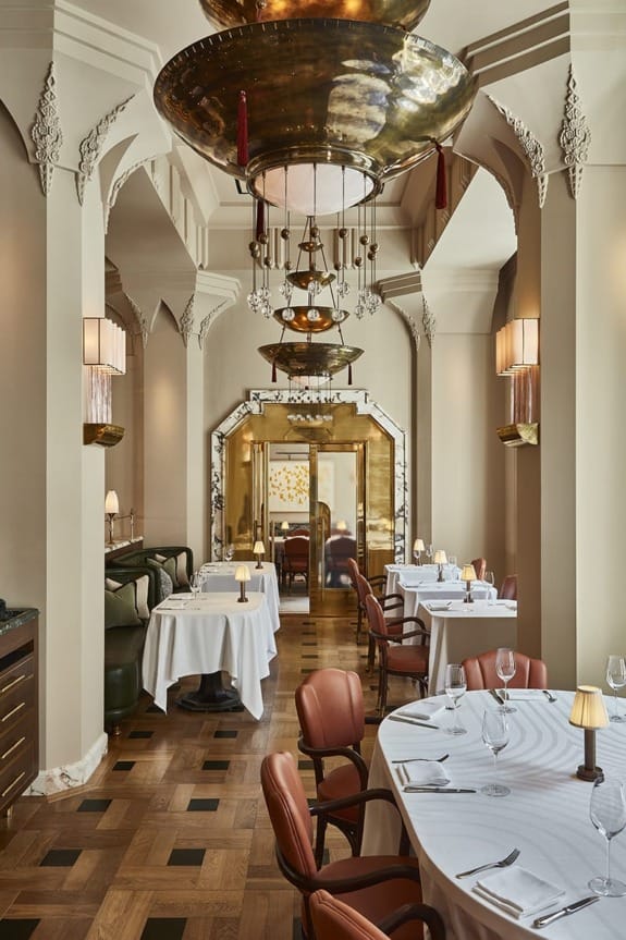 Elegant restaurant interior with ornate ceilings, round tables, white tablecloths, leather chairs, and soft lighting.