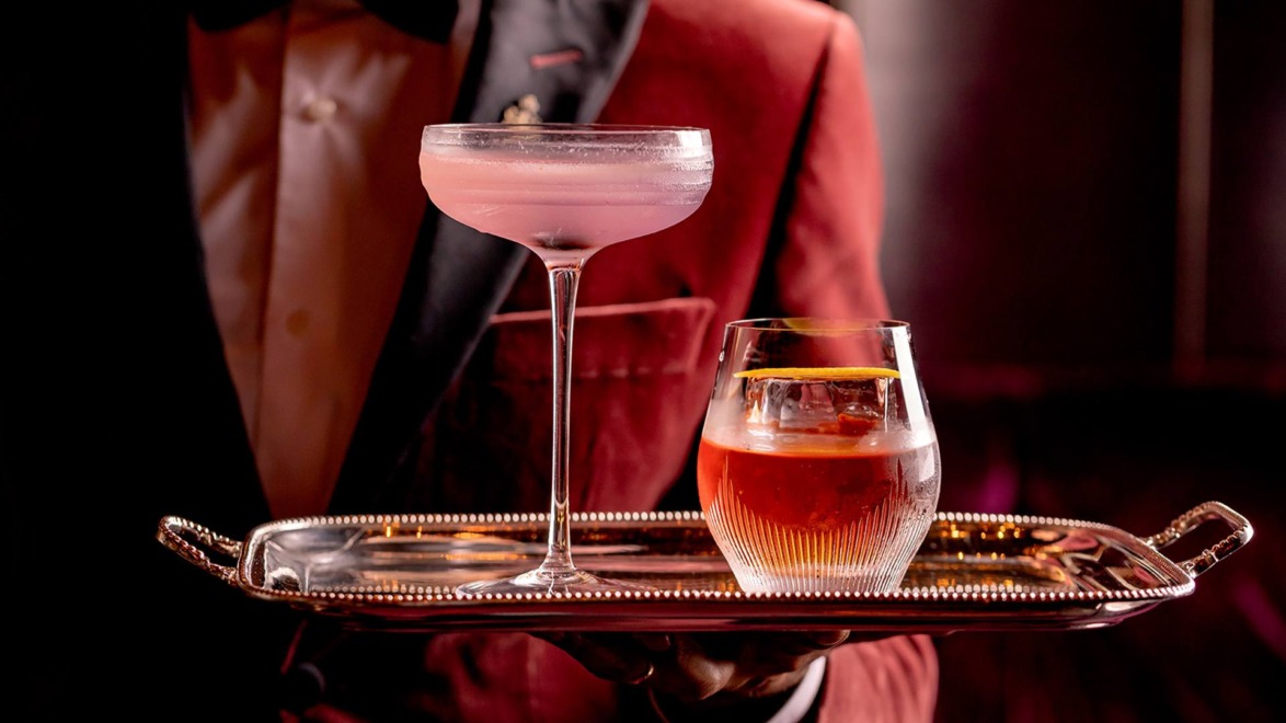 A bartender holding a tray with a pink cocktail in a coupe glass and a dark drink in a short glass.