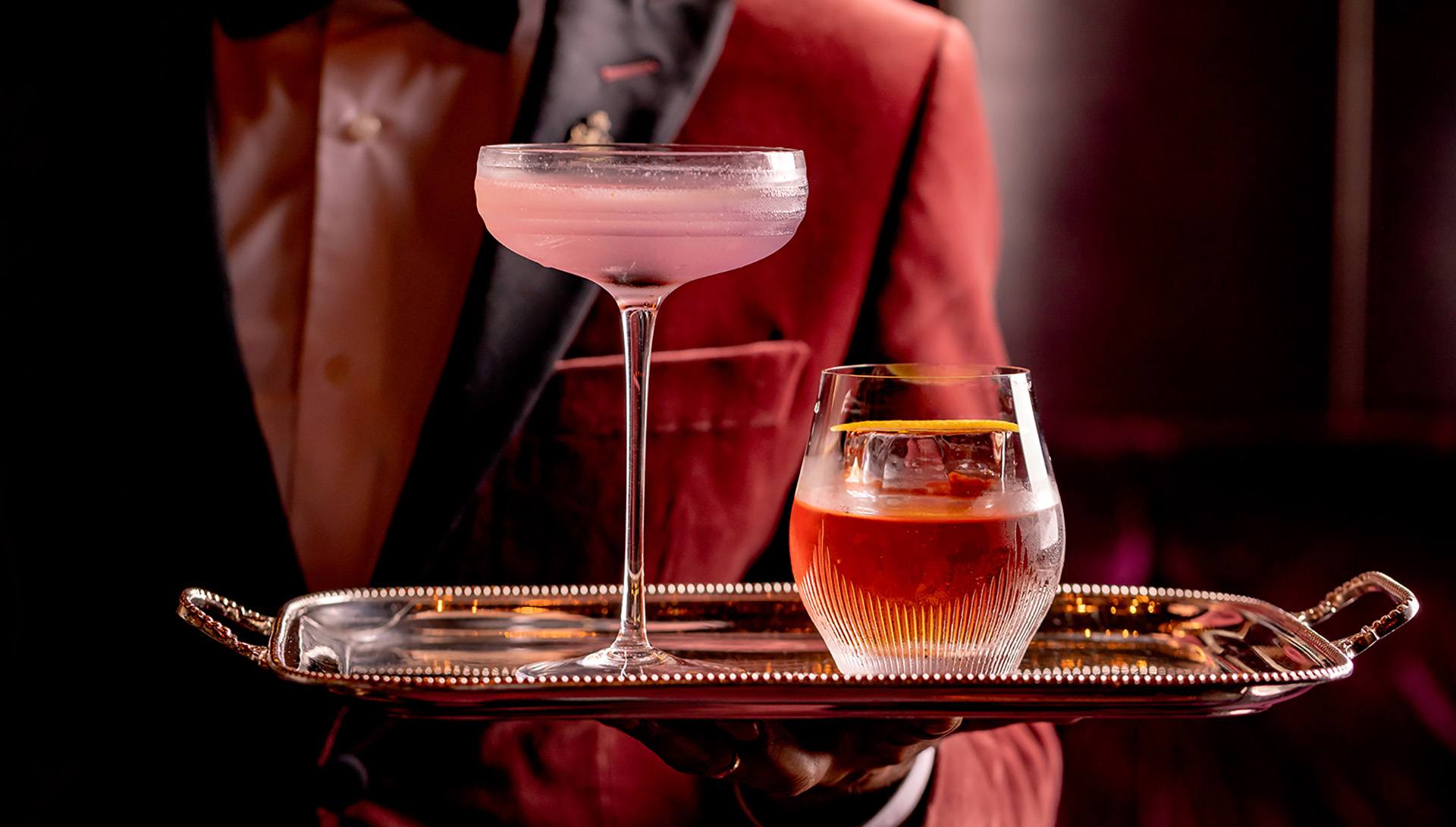 A bartender holding a tray with a pink cocktail in a coupe glass and a dark drink in a short glass.