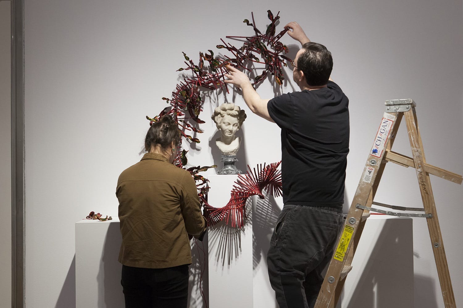 Two people assembling a red sculpture around a classical bust on a pedestal, with a ladder beside them.