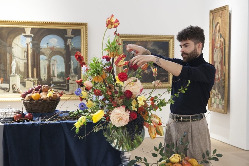 Man arranging colorful flower bouquet in art gallery setting with paintings and basket of fruit in the background.