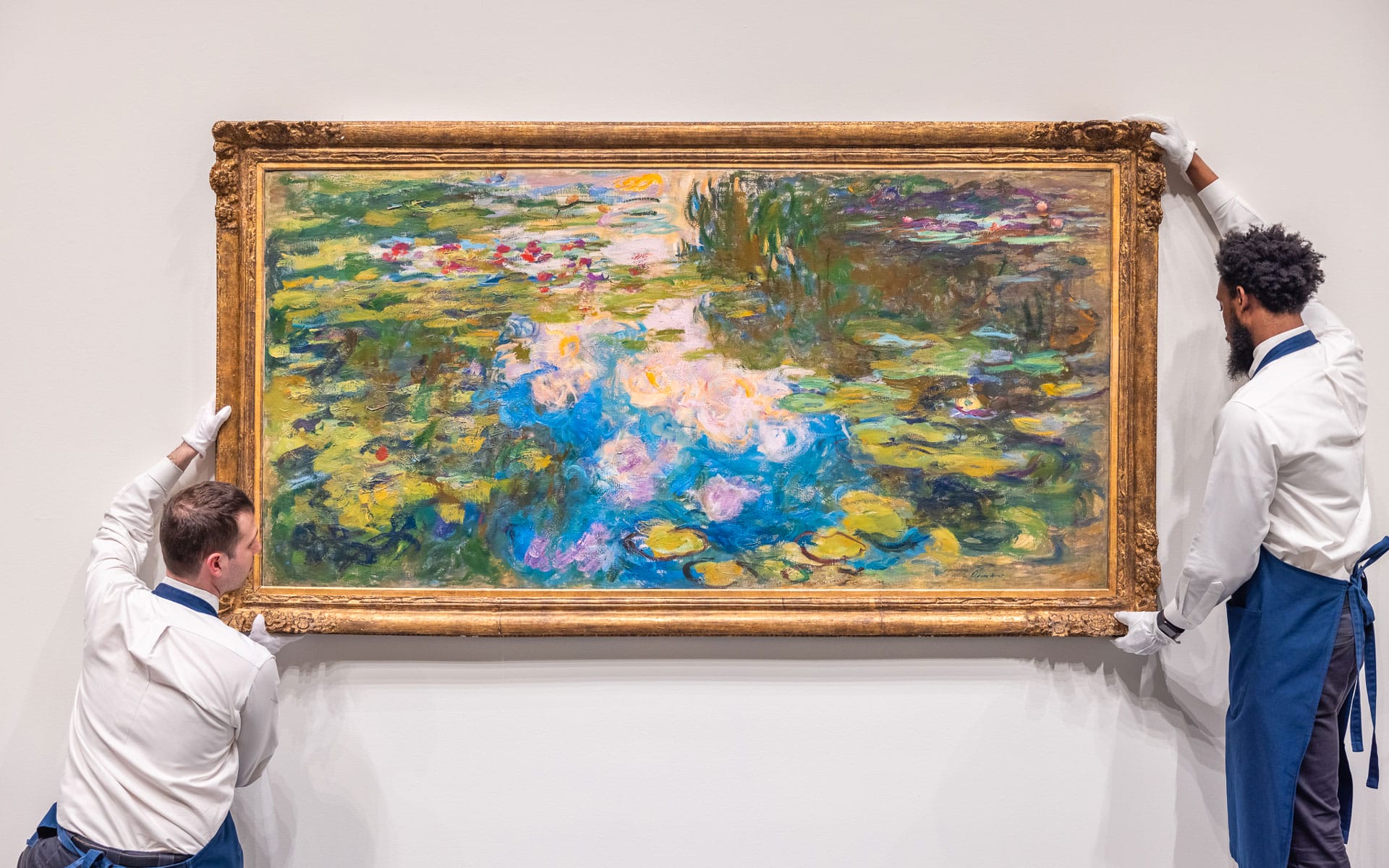 Two museum workers hanging a framed painting of a vibrant water lily pond on a gallery wall.