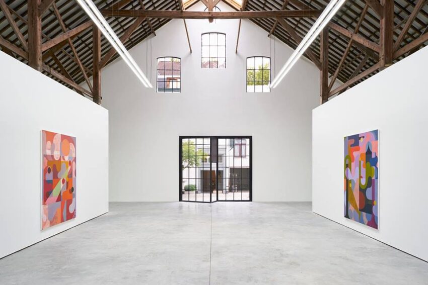 Contemporary art gallery with abstract paintings on white walls, high wooden ceiling, and large windows in a spacious room.