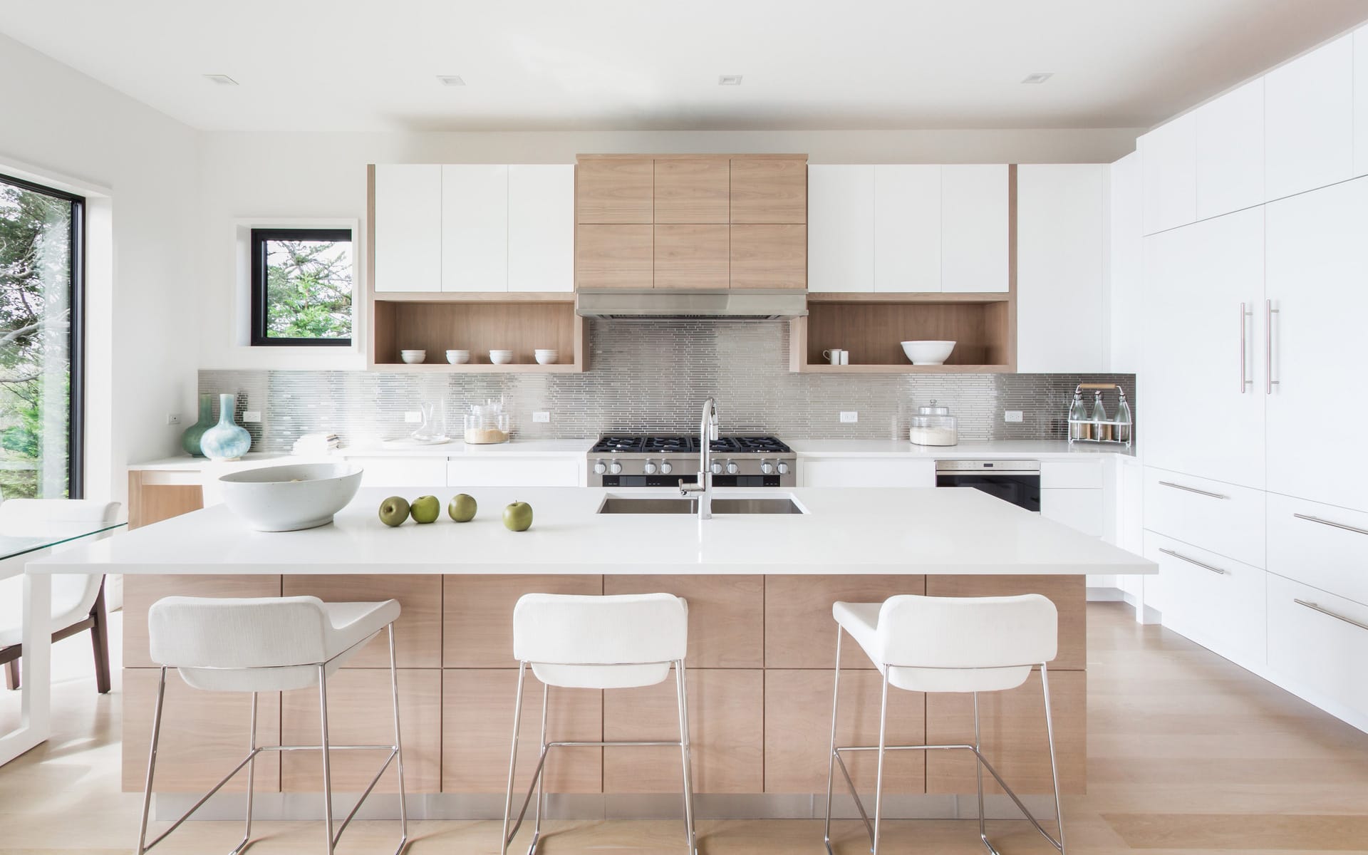 Modern kitchen with white cabinets, wooden accents, island with stools, stainless steel appliances, and large windows.