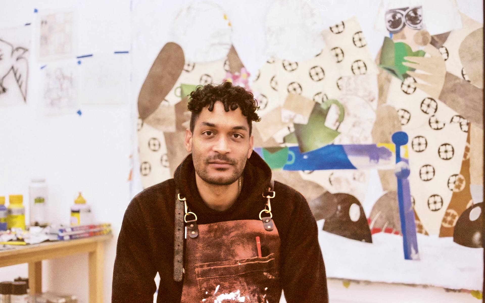 Man sitting in art studio, wearing an apron, with colorful abstract paintings in the background.