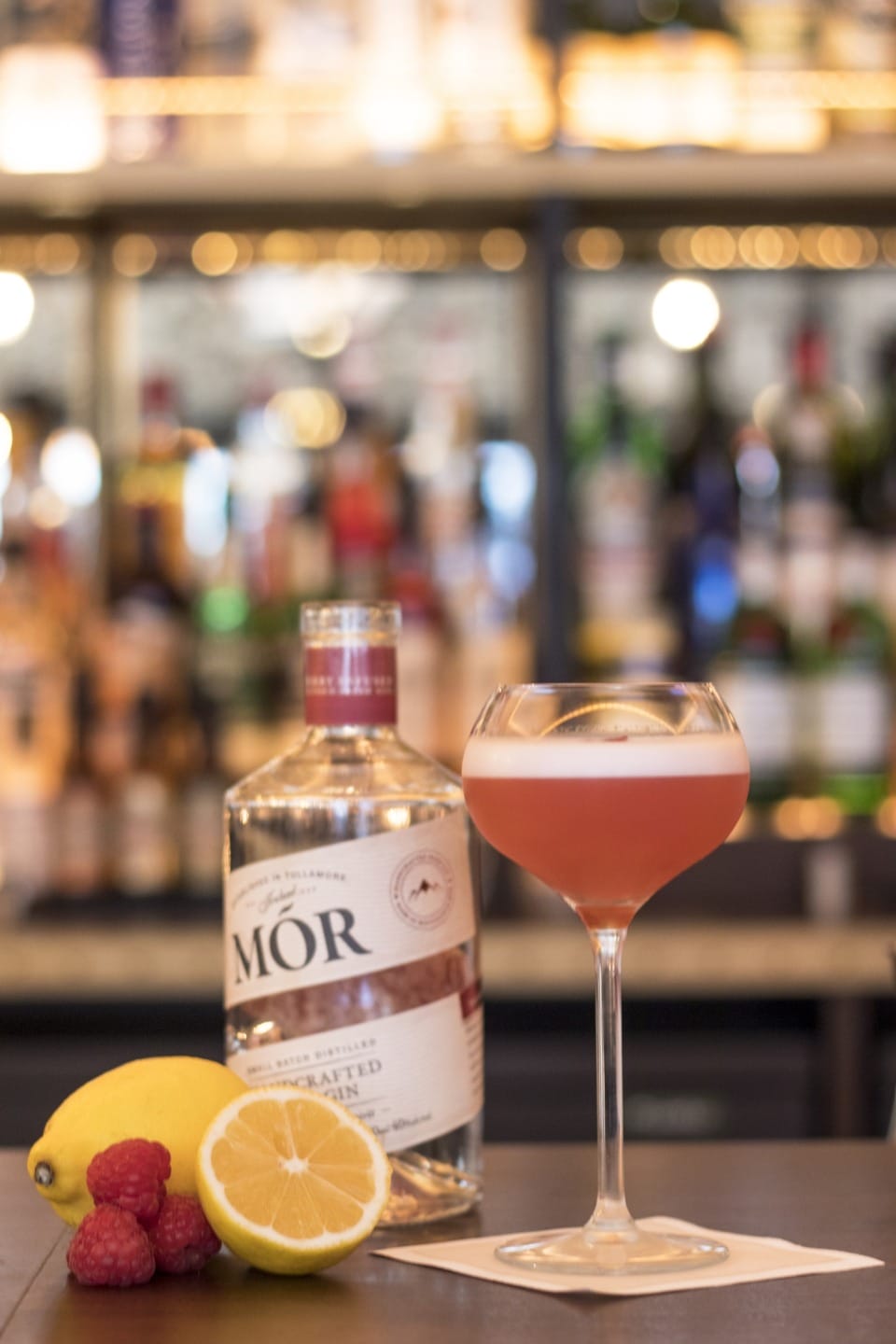 Cocktail in a stylish glass with a bottle of gin, surrounded by fresh lemon and raspberries on a bar counter.
