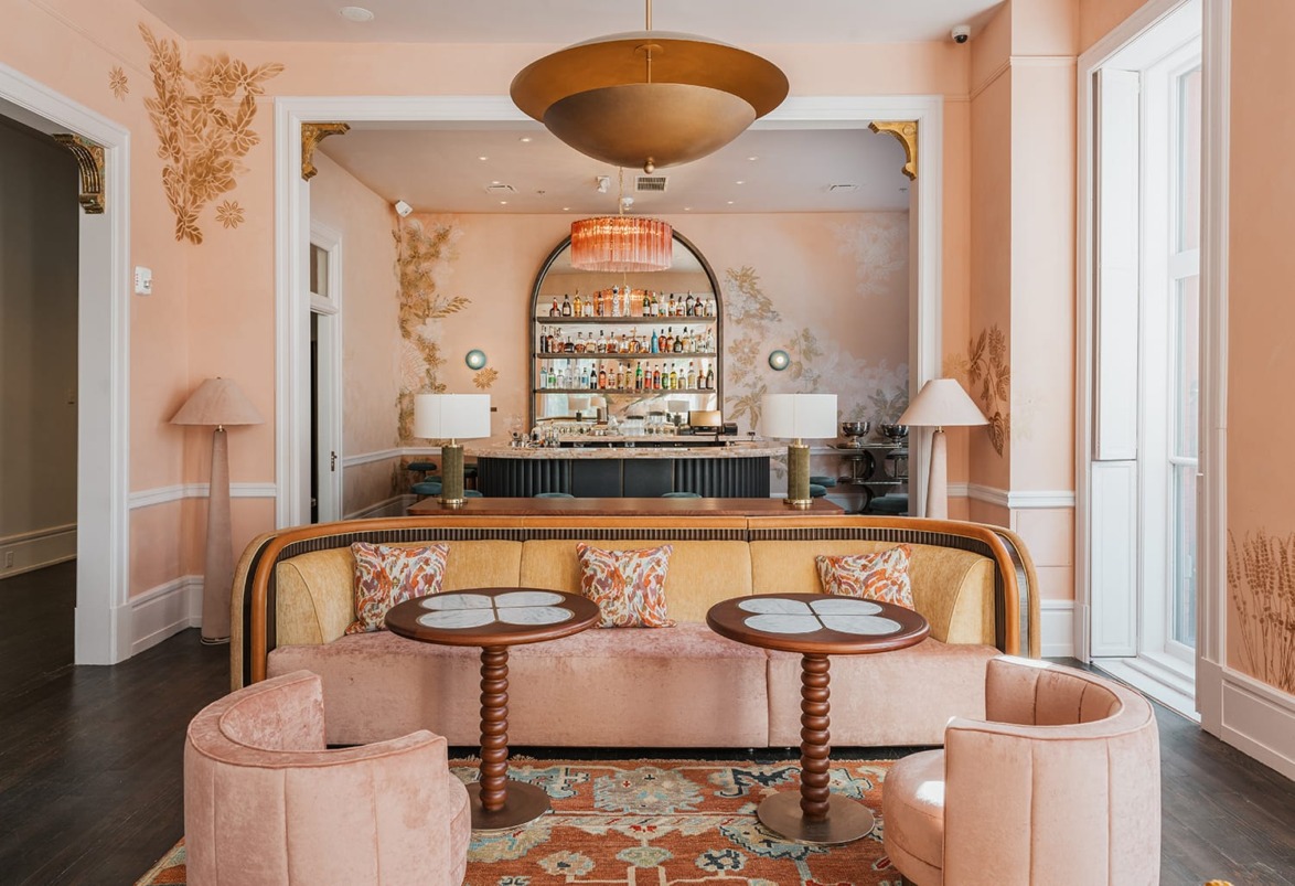 Elegant lounge with peach walls, velvet seating, stylish tables, large bar with assorted bottles, and ornate ceiling light.