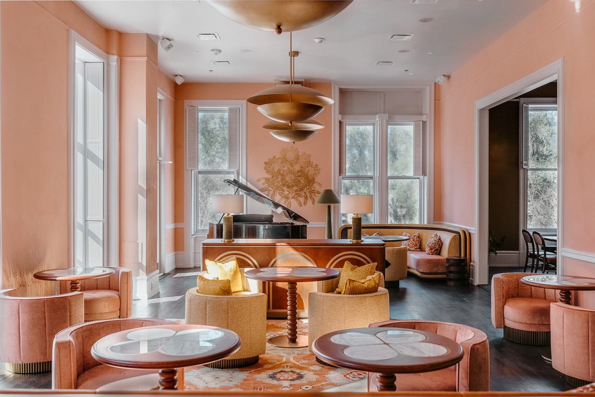 Elegant pink lounge with large windows, round tables, and cozy seating, featuring a grand piano and modern lighting.