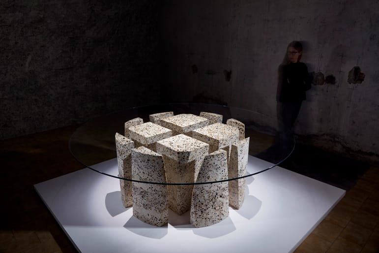 Modern glass table with geometric stone base in a dimly lit room, person standing in the background against a textured wall