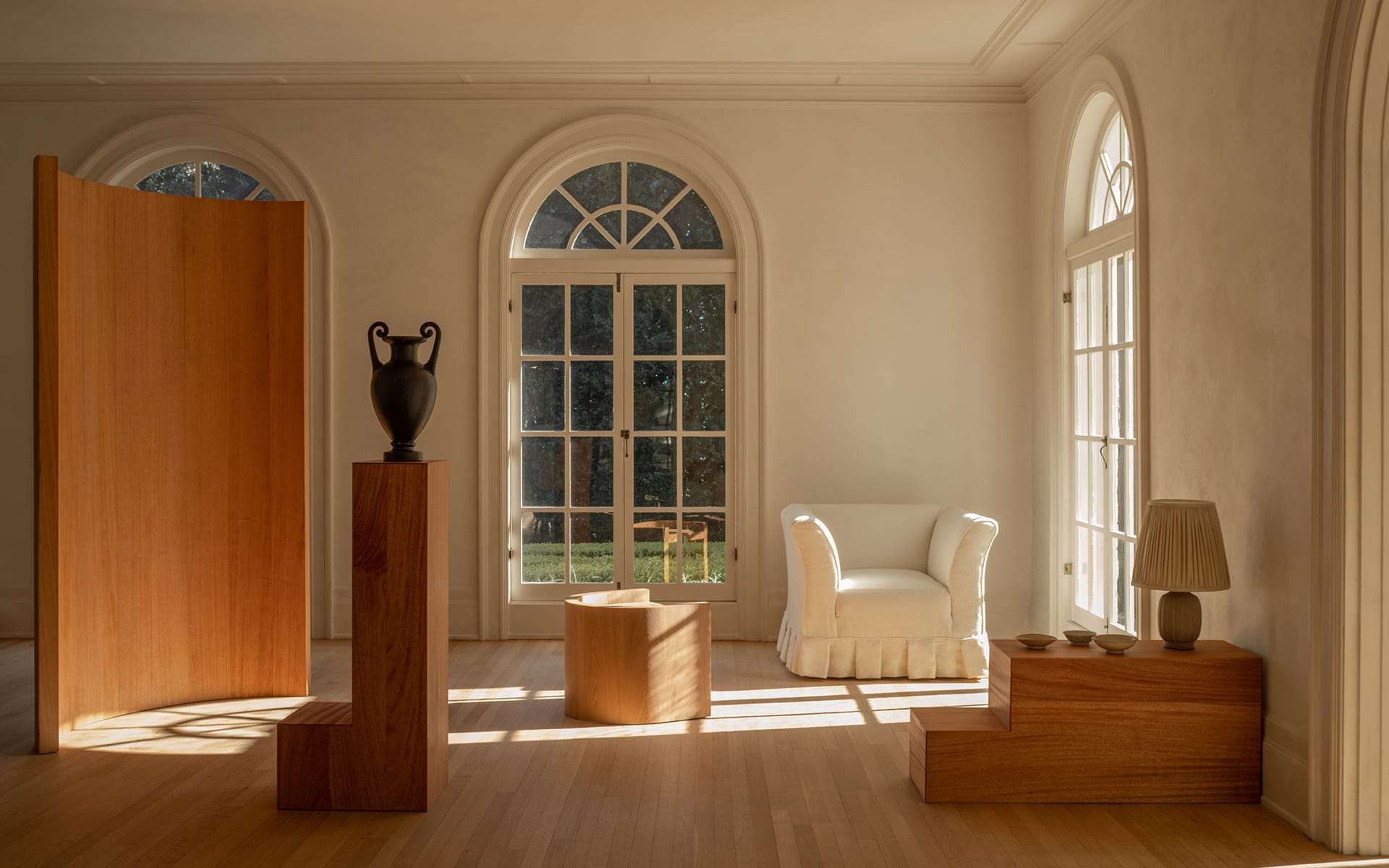 Minimalist living room with arched windows, wooden decor, sculpted black vase, white armchair, and soft lighting.