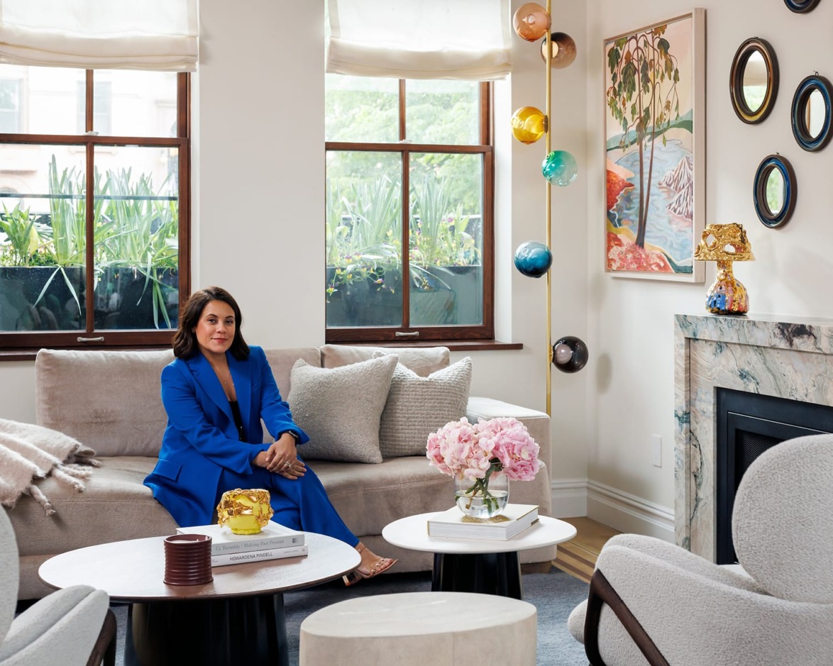 Person in blue suit sitting on a beige sofa in a stylish living room with large windows, modern decor, and plants outside.