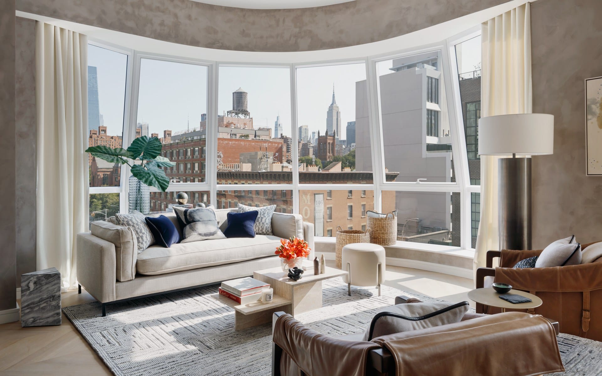 Modern living room with large windows, cityscape views, and stylish furniture including a beige sofa and leather armchairs.