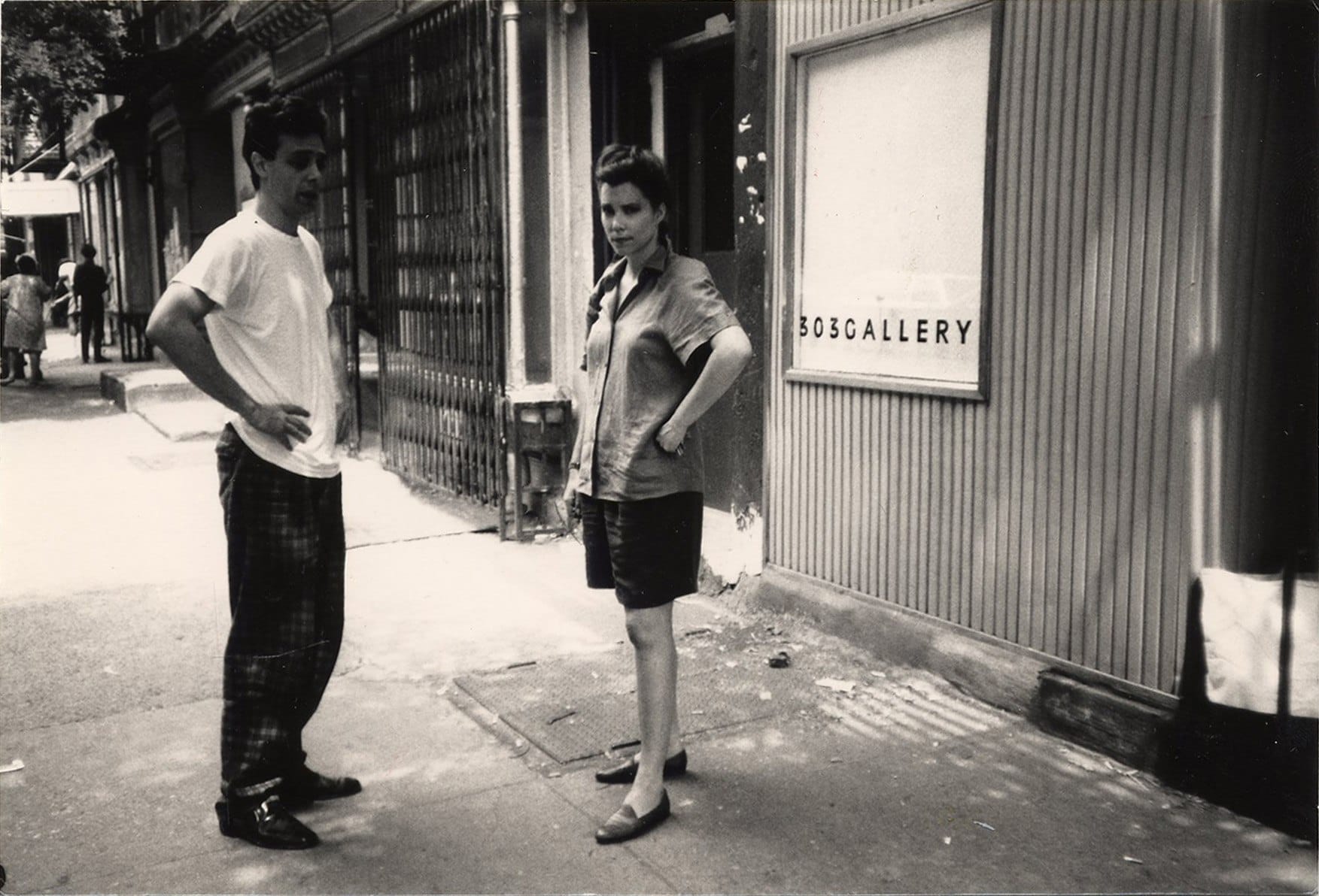 A man and woman stand on a sidewalk outside 303 Gallery in a black and white photo.
