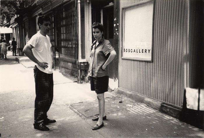 Two people standing on a sidewalk near a gallery entrance, one with hands on hips, urban street scene in black and white.