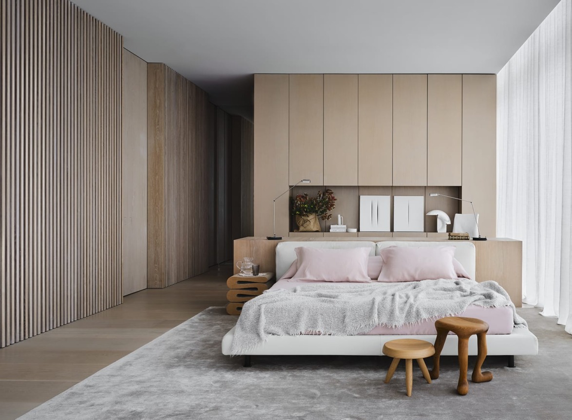 Modern bedroom with wooden accents, large bed with pink pillows, soft gray rug, and diffused natural light through sheer curtains