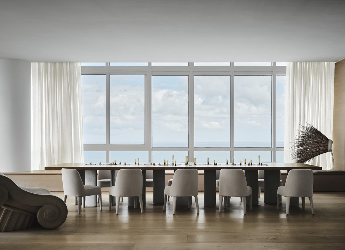 Modern dining room with a long table, surrounded by chairs, large windows, and ocean view in the background.