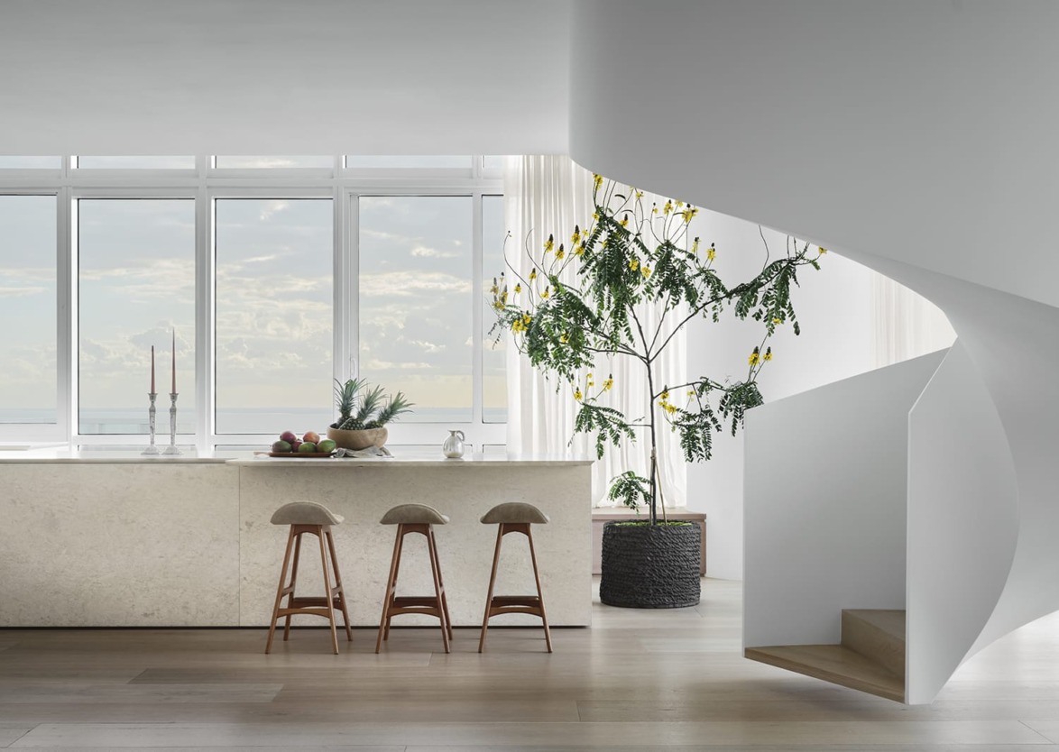 Modern minimalist kitchen with white island, three stools, large potted plant, and a spiral staircase by large windows.