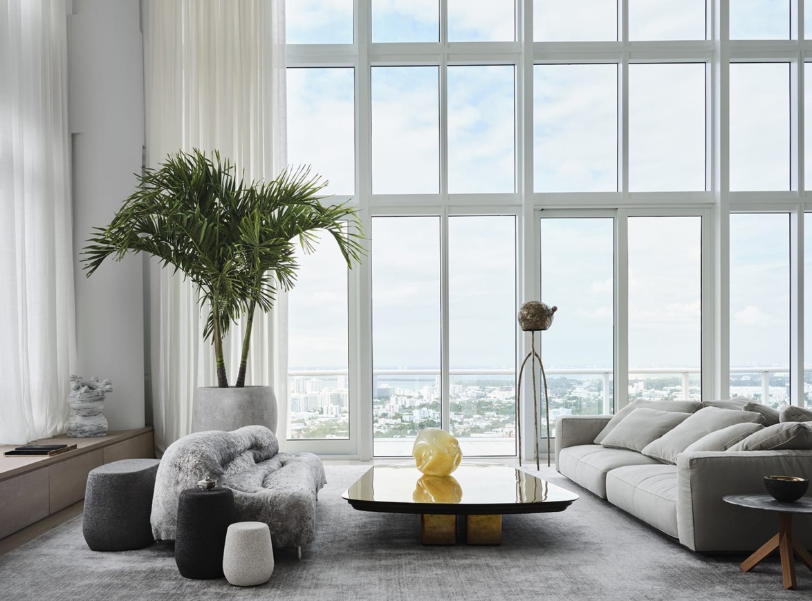 Modern living room with large windows, minimalistic furniture, a gray sofa, unique coffee table, and a tall indoor plant.