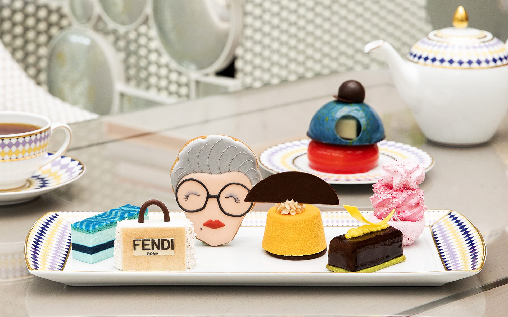 A selection of artfully designed desserts on a decorative plate with a teapot in the background.