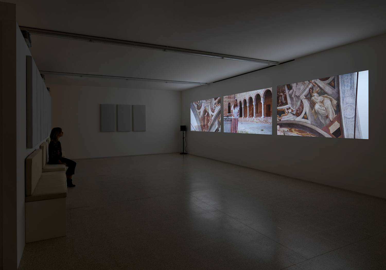 Person sitting in a dimly lit gallery observing three large illuminated images on the wall.