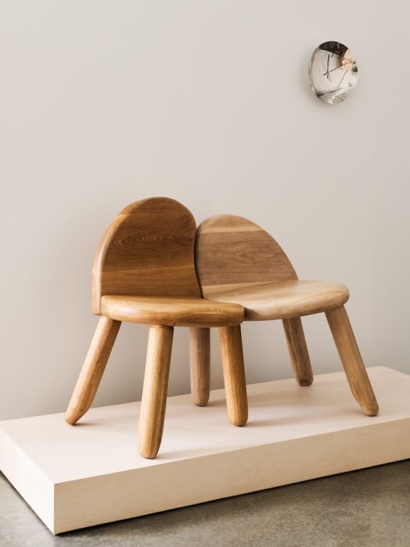 Two wooden chairs intertwined on a light-colored platform with a wall clock above them.