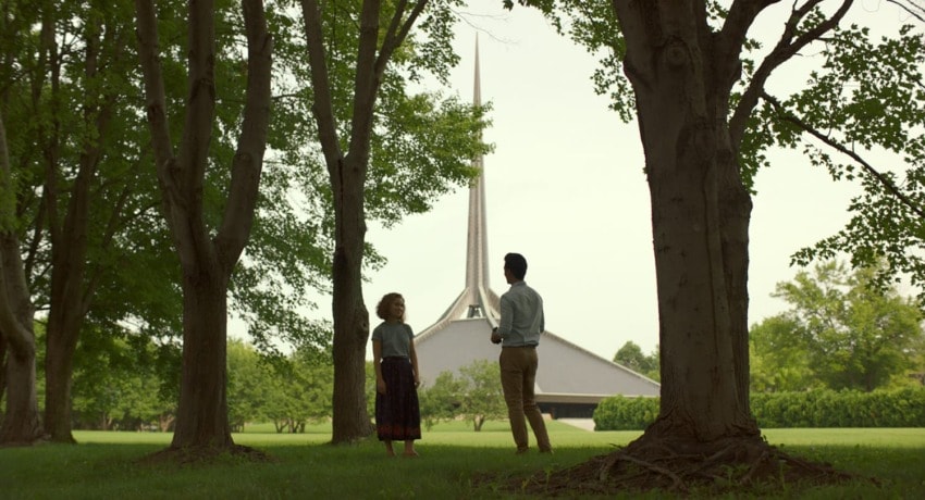 Two people standing in a park-like setting with trees, talking near a building with a tall, pointed spire in the distance.