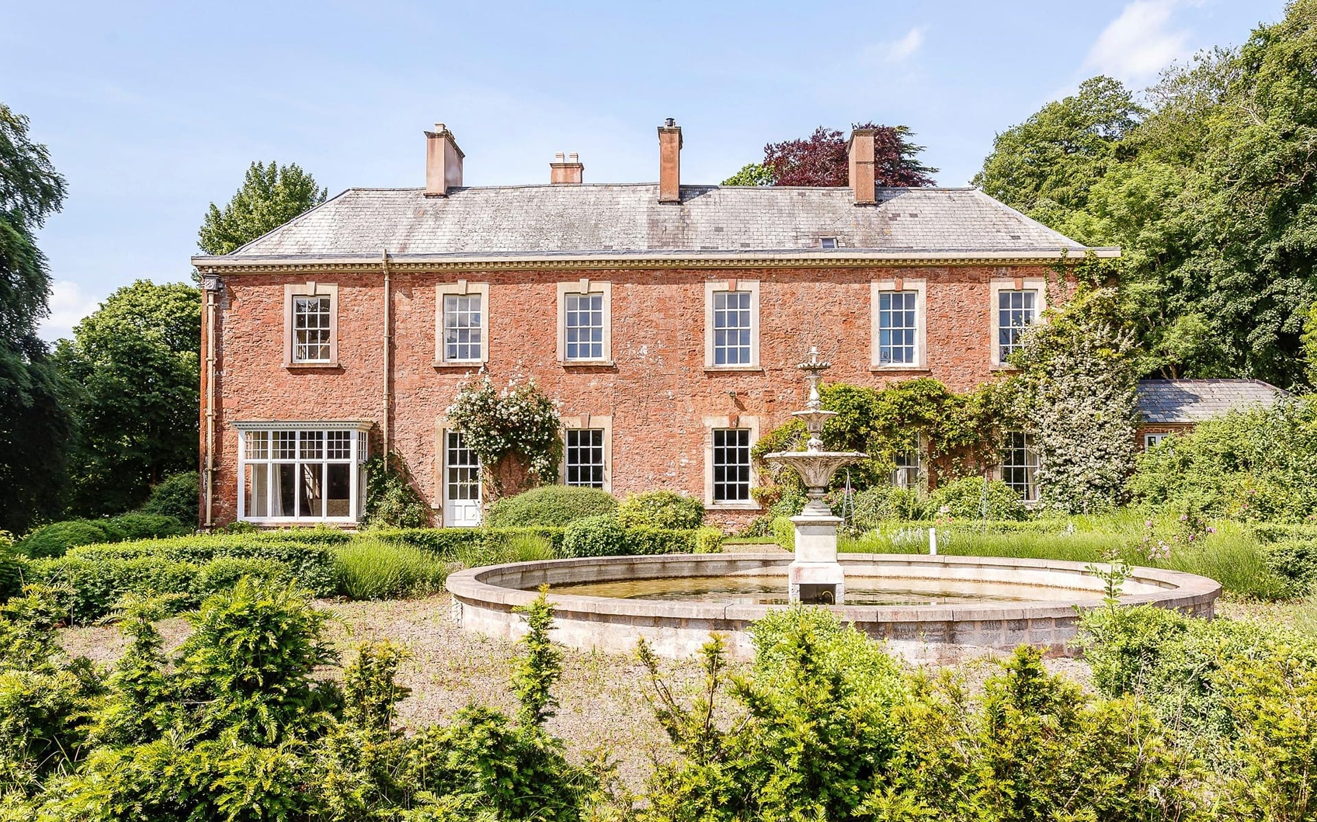Large brick mansion with a fountain in front, surrounded by lush greenery and under a clear blue sky.