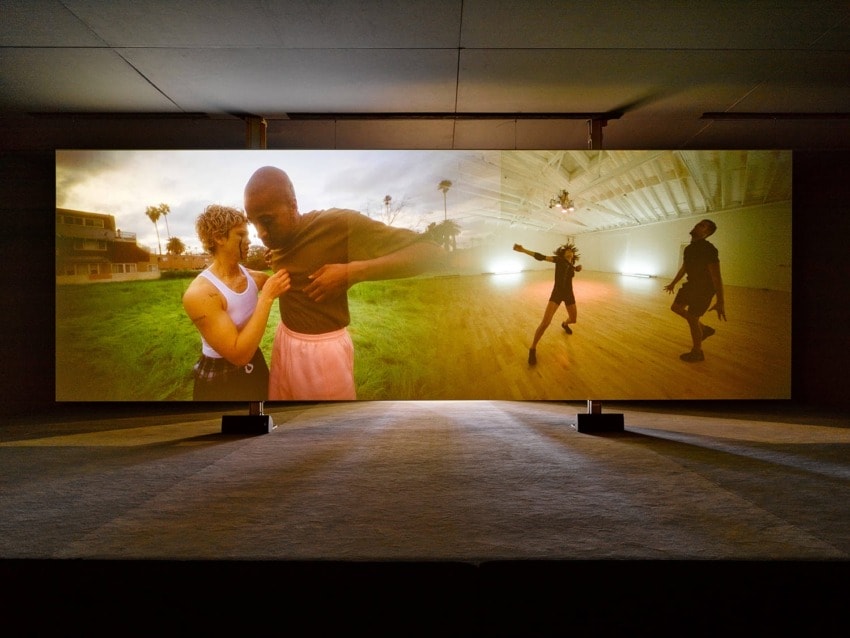 Two large screens displaying dancers performing in an indoor studio and an outdoor setting with a sunset backdrop.