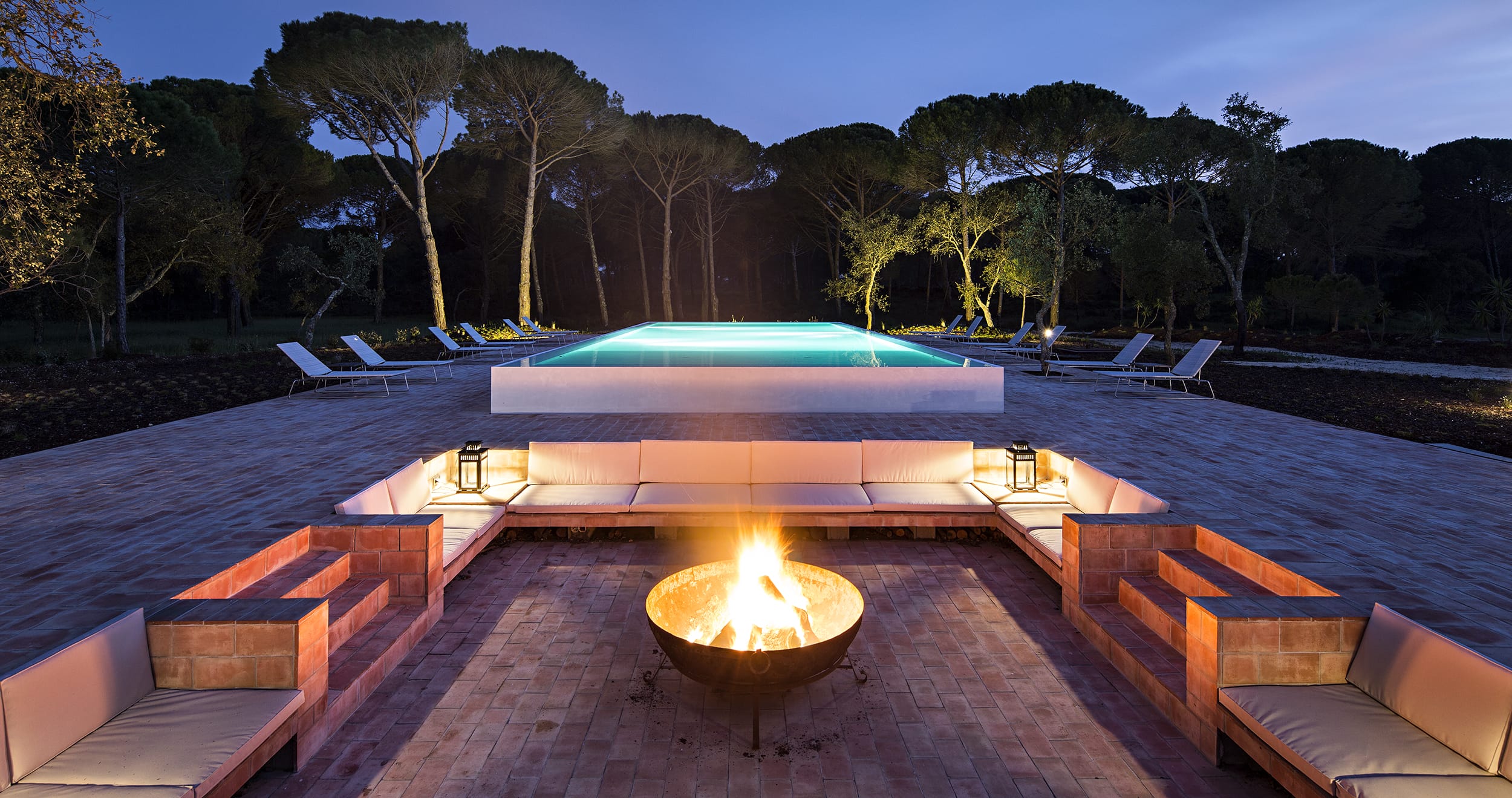 Outdoor seating area with fire pit overlooks an illuminated pool surrounded by trees at dusk, creating a tranquil scene.