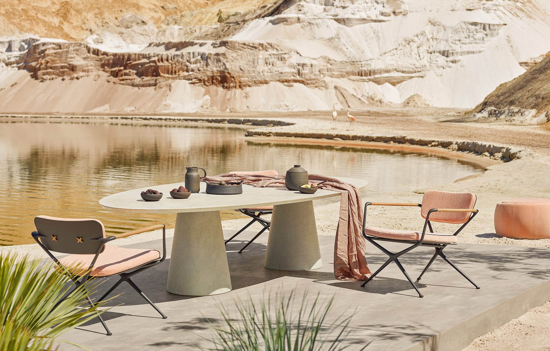Outdoor modern dining set by a lake with a concrete table, two chairs, and a teapot, surrounded by a desert landscape.