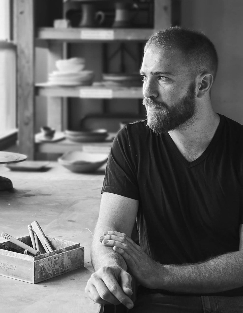 Man sitting in pottery studio, gazing thoughtfully, with pottery tools and shelves in the background, black and white image.