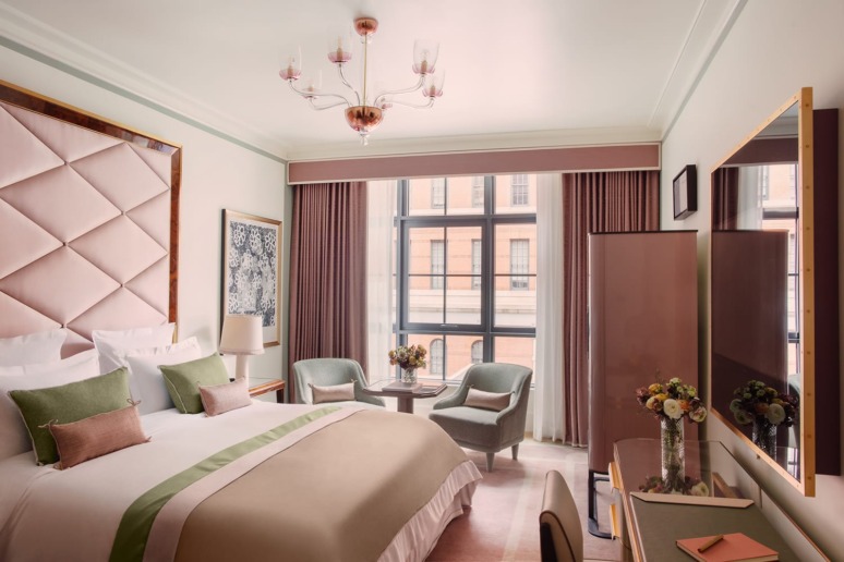 Elegant hotel room with a large bed, cushioned headboard, two armchairs, a small table, large window, and soft lighting.