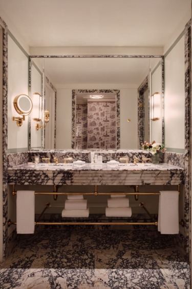 Elegant marble bathroom with floral wallpaper, large mirror, twin sinks, gold fixtures, and neatly folded towels.