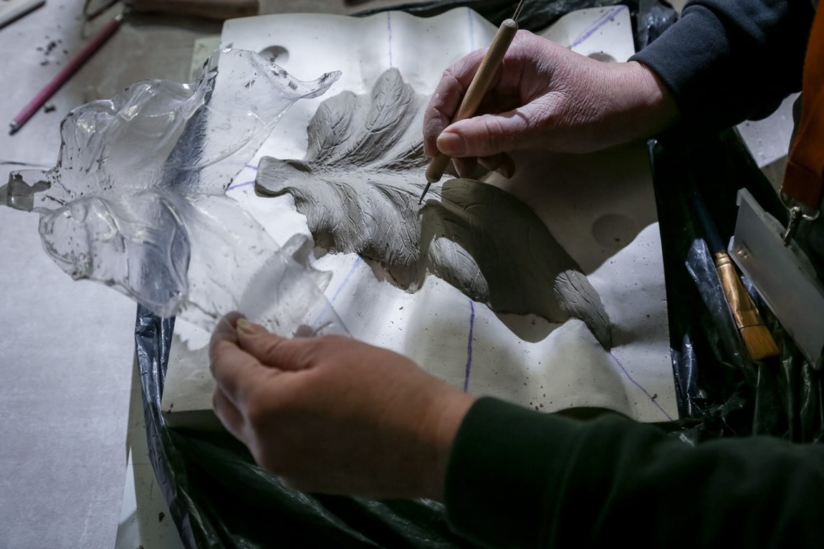 Person sculpting floral design in clay with a carving tool, holding a glass leaf for reference.