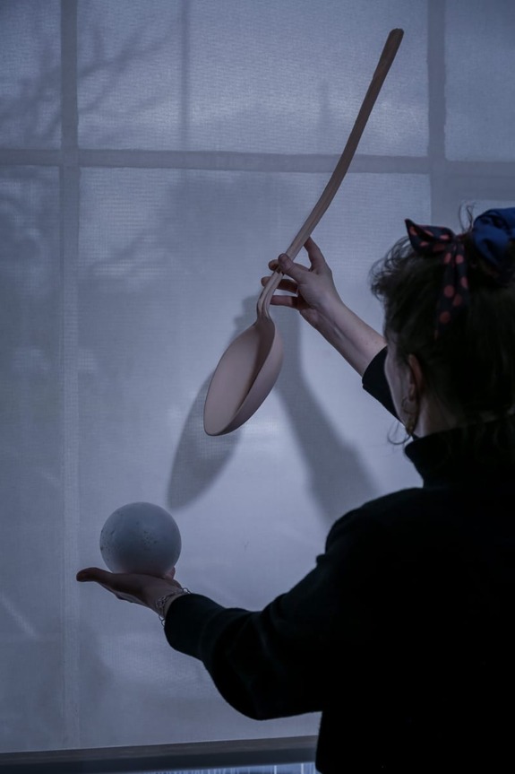 Person holding a large wooden spoon in one hand and a gray sphere in the other against a backlit window.